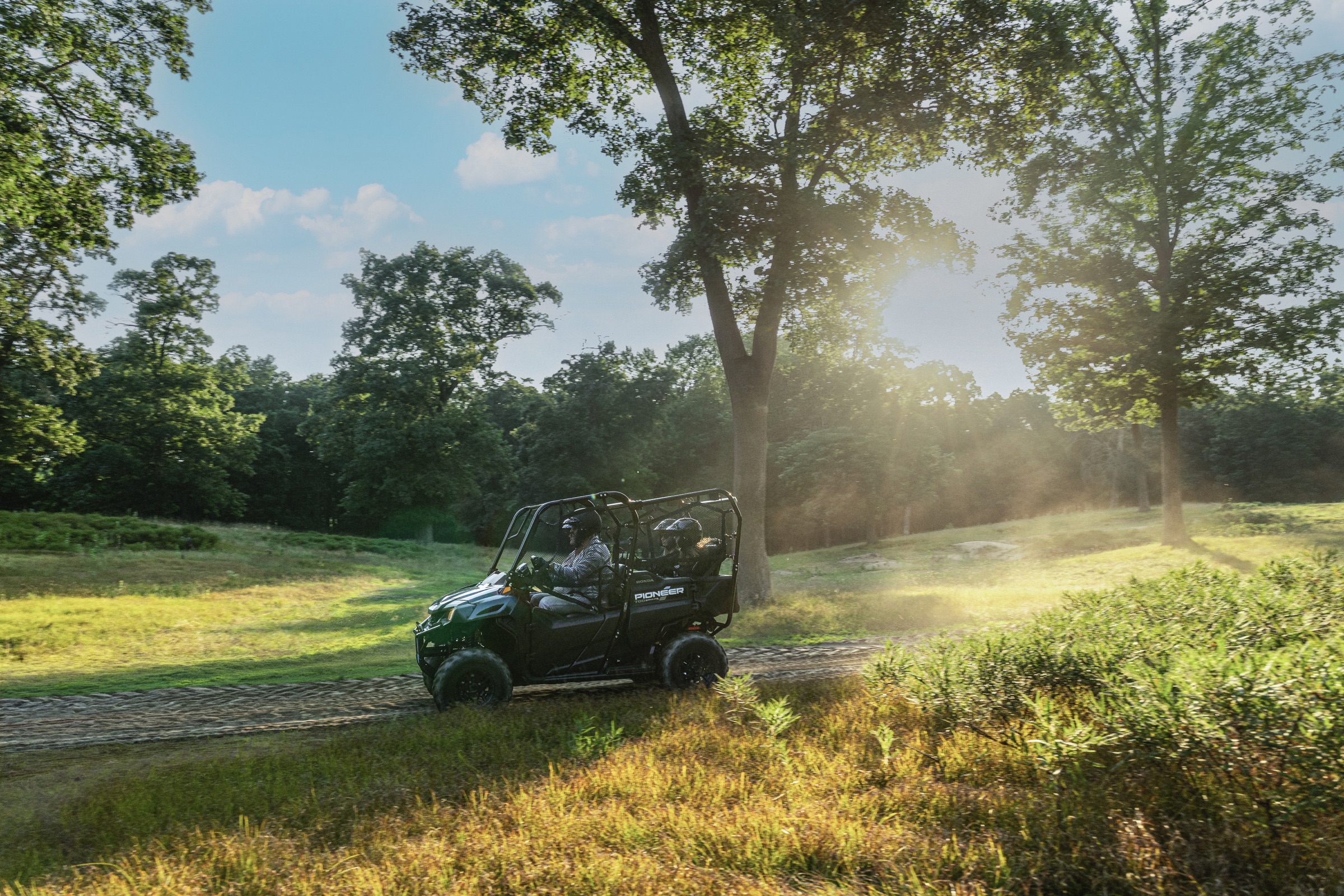 Two helmeted occupants riding a Honda Pioneer side by side on a wooded trail